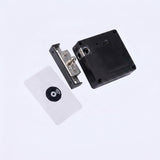 A close-up studio shot of a small electronic lock or door control component floating against a light gray background. The product appears to consist of a black rectangular housing, a silver metal latch or bracket piece, and a white rectangular card with a black circular symbol.